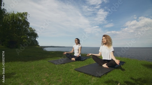 Two women meditating outdoors seated on the edge of coast near the sea, lotus pose folded fingers mudra gesture do yoga exercise in morning.