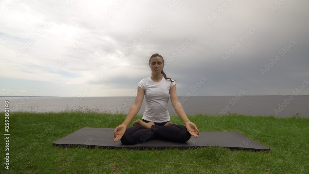 Young fit woman practice yoga on coast near the lake or sea. Woman sitting in Knee Pile pose and meditating