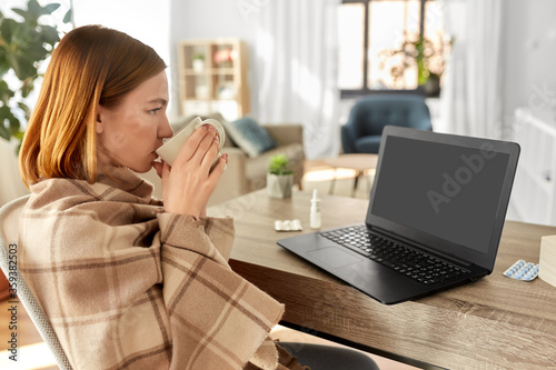 healthcare, technology and people concept - sick young woman in blanket drinking tea and having video call on laptop computer at home