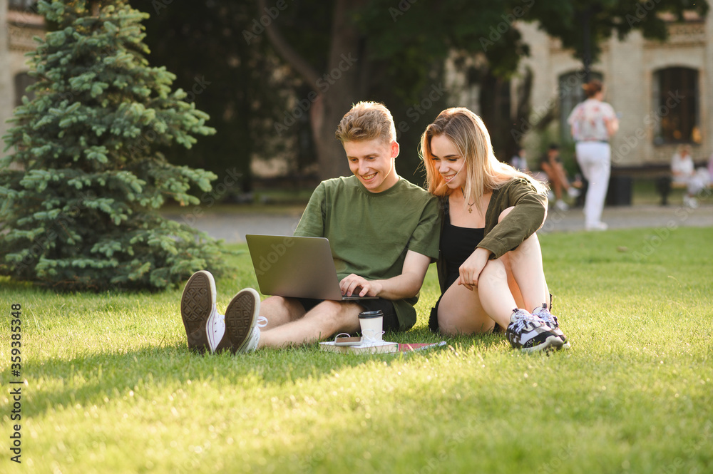 Couple of happy students using laptop while sitting on university campus lawn, looking at screen and smiling. Smiling girl and young man studying with laptop at park break. Student leisure. Couple of happy students using laptop while sitting on university campus lawn, looking at screen and smiling. Smiling girl and young man studying with laptop at park break. Student leisure.