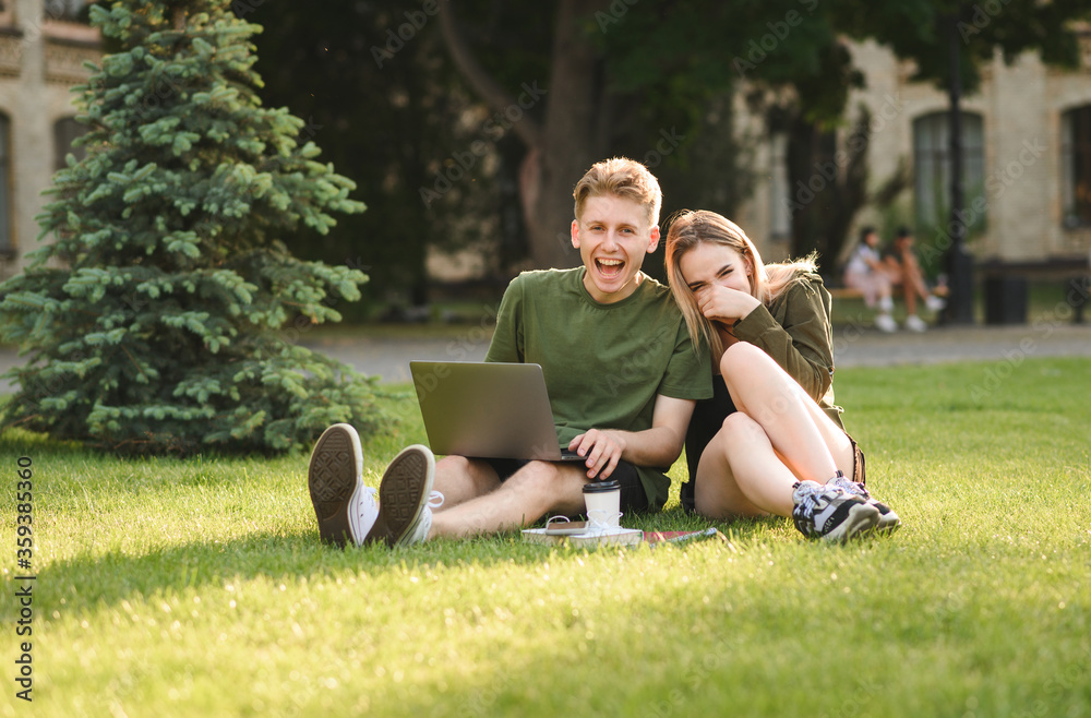 Couple of smiling students guy and girl sitting on grass in university park with laptop, looking at camera and laughing. Joyful girl and young guy having fun after lessons on the lawn Couple of smiling students guy and girl sitting on grass in university park with laptop, looking at camera and laughing. Joyful girl and young guy having fun after lessons on the lawn