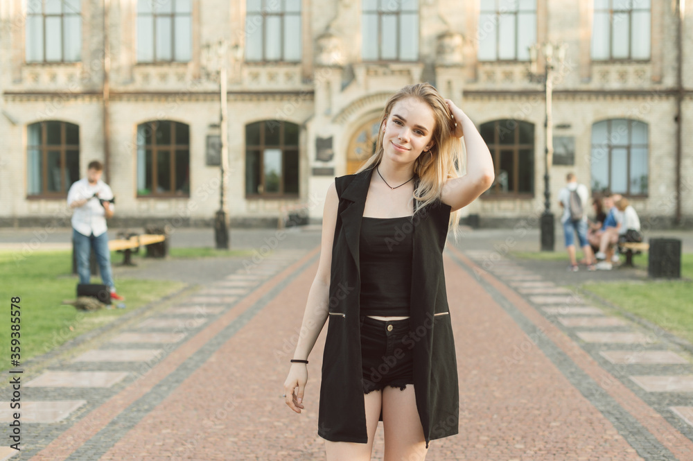 Happy lady in black casual clothes is standing on the door on a university background and posing at camera. Cute girl stands on the background of the old building and looks in camera with a smile