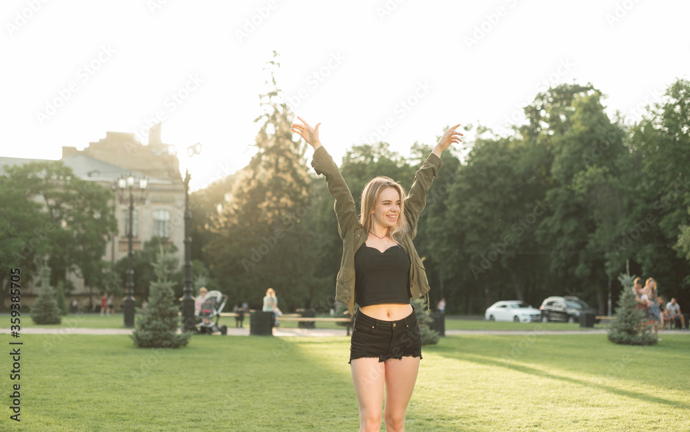 A happy girl in short shorts stands in the park and rejoices with her hands raised, looking away with a smile on her face. Portrait of joyful lady rejoicing in victory in park on lawn.