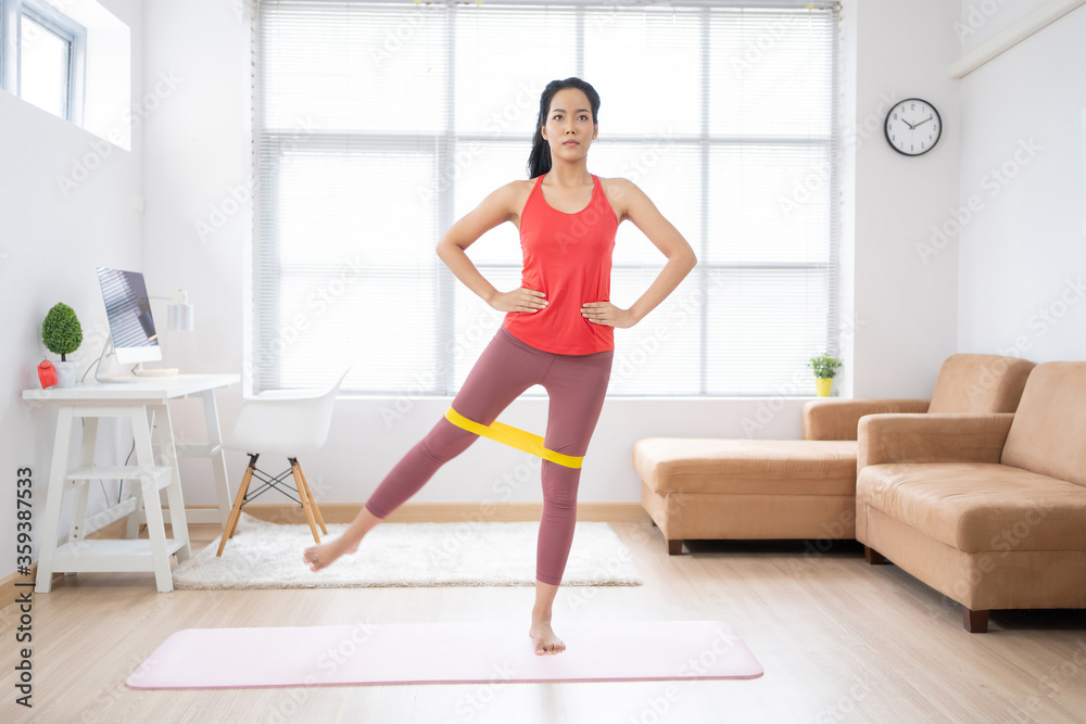 Asian woman exercising at home on a yoga mat. She uses rubber resistance.