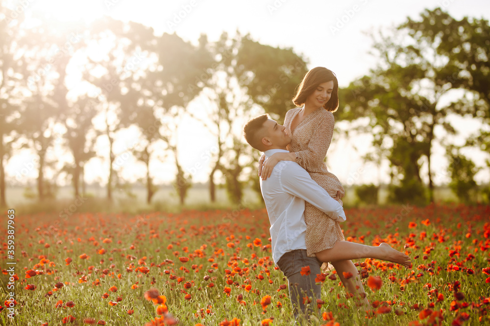 Fototapeta premium Romantic couple having fun on the poppy field on the sunset. A boy holds a girl on his hands and being happy among red poppy flowers. Peace, love, family value, togetherness concept.