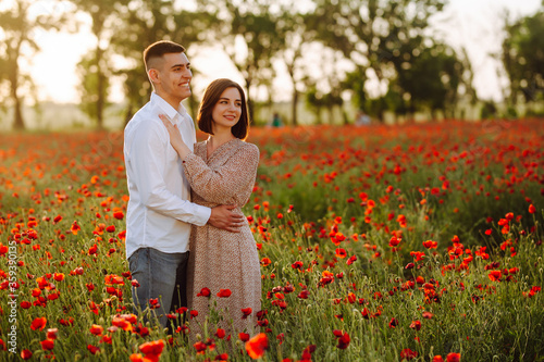 Young romantic loving married couple is standing among the red poppy flowers on the field on the beautiful sunset. Attractive boy and tender girl hug and kiss feeling happy. Love, family concept.