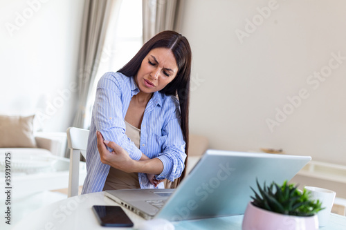 Portrait of young stressed woman sitting at home office desk in front of laptop, touching aching elbow with pained expression, suffering from elbow after working on laptop © Graphicroyalty