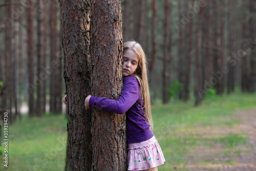 Blond girl with closed eyes huggies tree in the pine forest, enjoying nature. Love nature and care of nature concept.