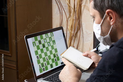 A man in a medical mask solves a chess problem online. Chess board with figures on a laptop screen. Leisure in self-isolation. International chess day. photo