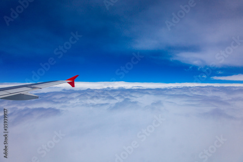 The wing of the plane flies over white clouds, the background is blue sky, view through the window.