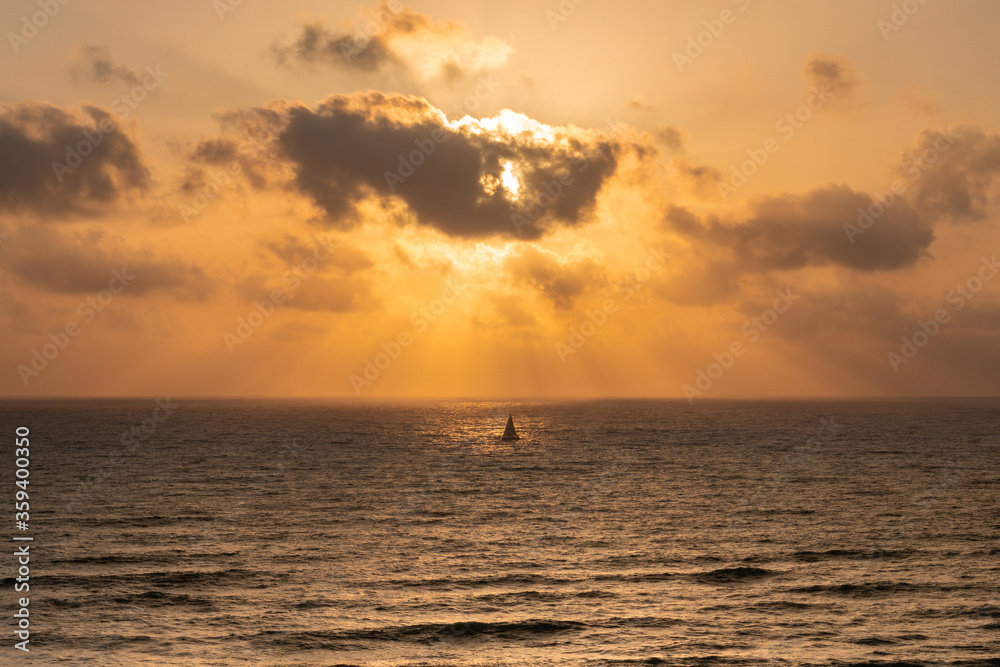 Beautiful sunset with golden colors over the sea with clouds covering/hiding the sun, the sunbeam and sun rays are lighting the boat in the middle of the ocean, reflecting off the sun on the water