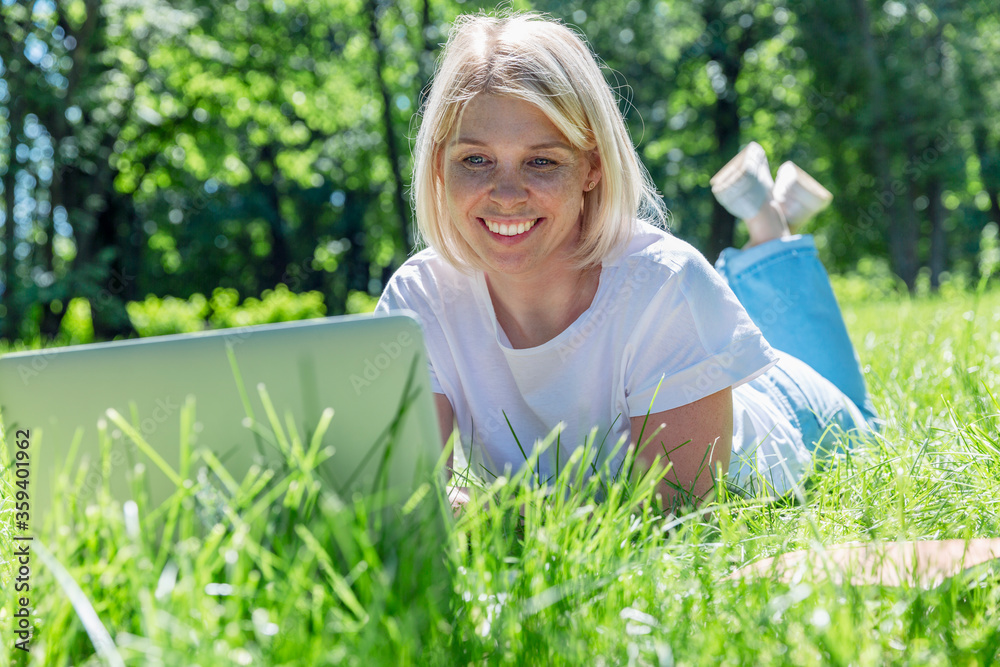 A smiling young blonde woman lies on the grass in a park with a laptop on a summer sunny day. Blogging, online communication, distance work and training. A smiling young blonde woman lies on the grass in a park with a laptop on a summer sunny day. Blogging, online communication, distance work and training.