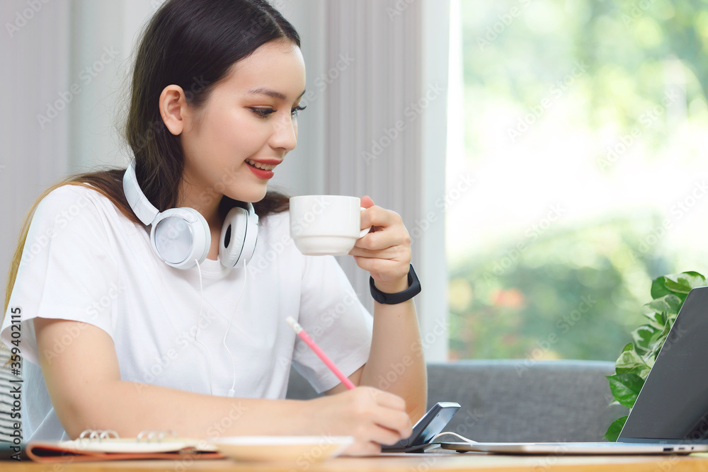Asian woman student sitting at the table in living room, drinking coffee and using computer laptop when studying online course