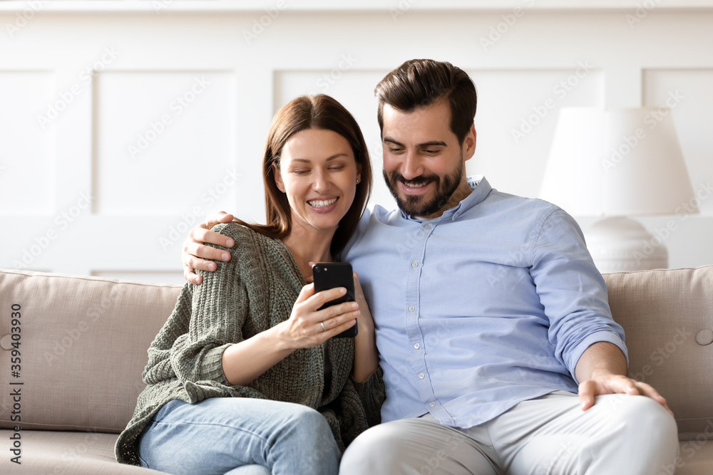 Smiling man and woman hugging, using smartphone together, sitting on cozy couch at home, young couple having fun with mobile device, looking at screen, watching video, chatting or shopping online