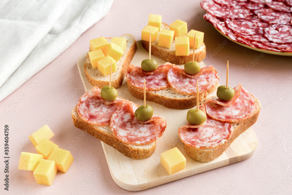 Sandwiches of smoked sausage and white bread on a pink paper background. The dish is decorated with green olives. There is a plate of sliced sausage on the back.