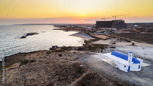 Aerial bird's eye view of coastline sunset, traditional landmark white and blue chapel at Agia Thekla beach, Ayia Napa, Famagusta, Cyprus from above.Tourist attraction golden sand bay, church, sunbeds photo