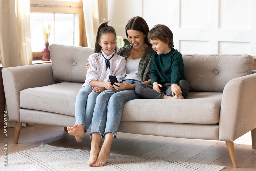 Smiling mother with son and daughter using smartphone, happy mum with adorable children relaxing on cozy couch, shopping or chatting online, watching video, looking at phone screen, enjoying weekend