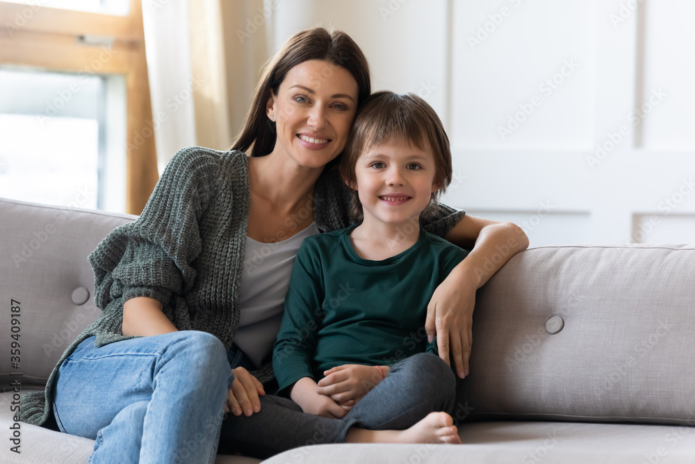 Family portrait smiling mother hugging embracing little son, sitting on couch, looking at camera, happy mum with preschool child kid posing for photo at home together, enjoying leisure time