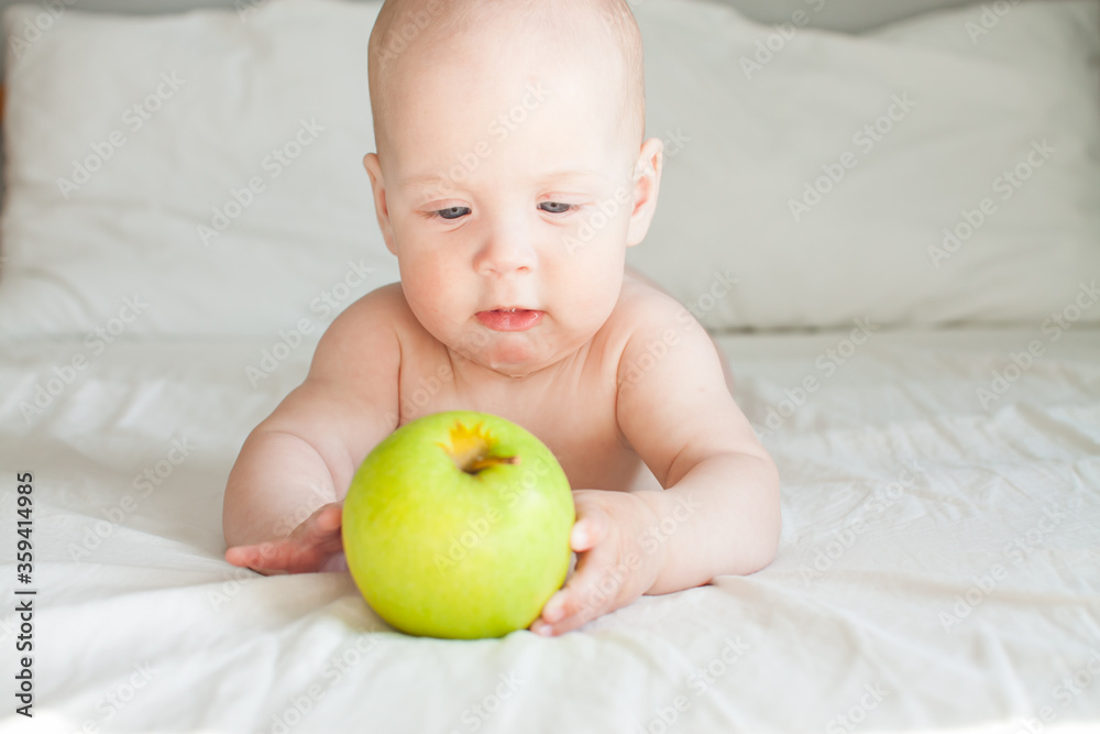 Cute cheerful baby lies on a white bed and looks at a green apple lying in front of her Cute cheerful baby lies on a white bed and looks at a green apple lying in front of her