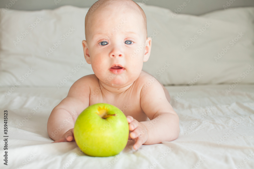 Cute cheerful baby lies on a white bed and looks at a green apple lying in front of her