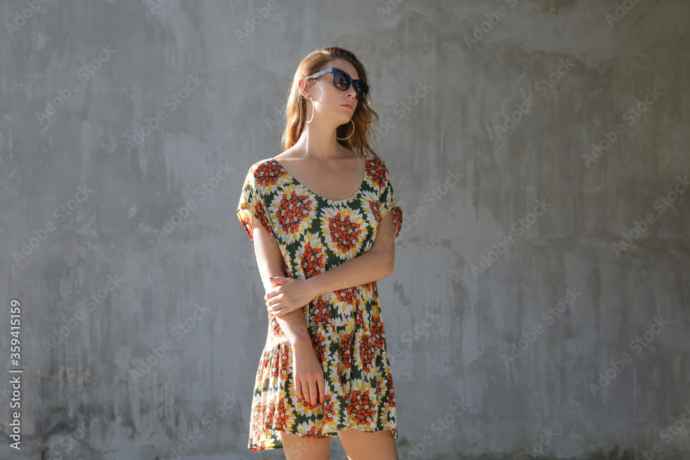 Fototapeta premium Outdoor portrait of stylish fashionable woman in colorful dress in front of a grey wall. Casual summertime hippie boho outfit, spring-summer fashion trend.
