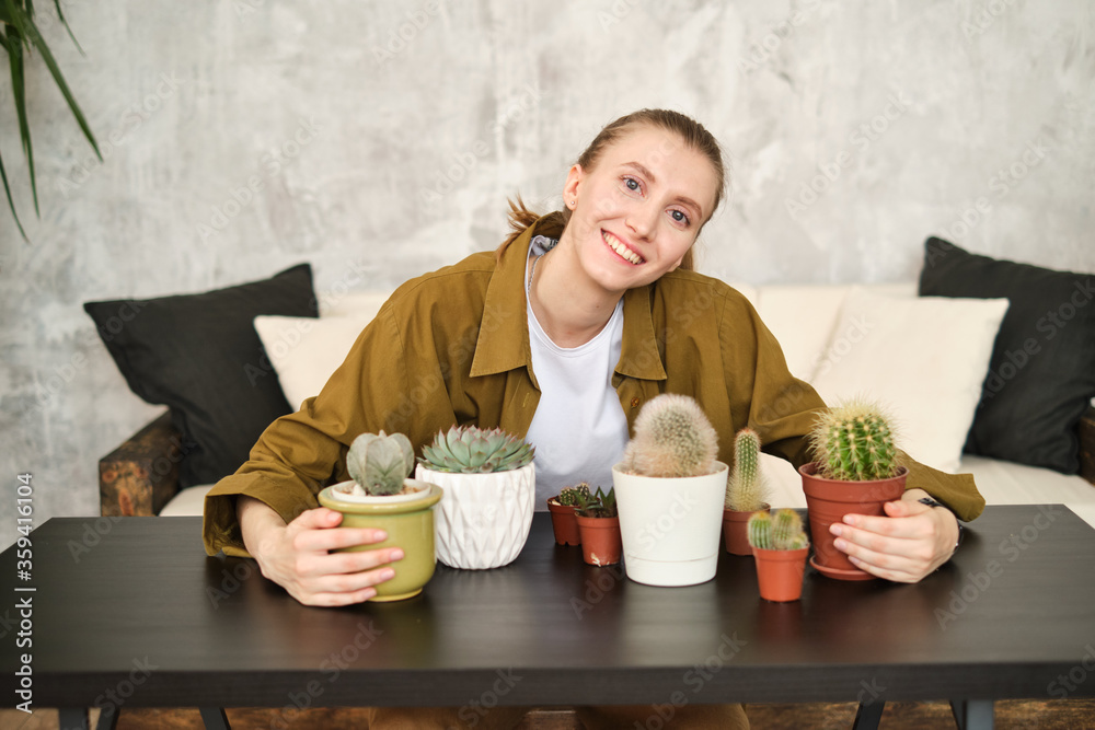 Portrait of smiling young woman taking care of plants and home flowers. Home gardening and slow life rituals. On the table are pots with cactus and succulents. Likes gardening and plants. Portrait of smiling young woman taking care of plants and home flowers. Home gardening and slow life rituals. On the table are pots with cactus and succulents. Likes gardening and plants.