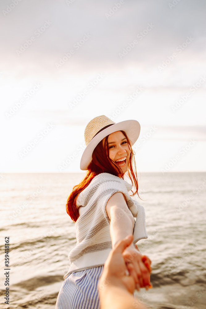 Smiling long haired woman in a hat holding boyfriends hand along empty ocean beach sand at sunset . Follow me concept