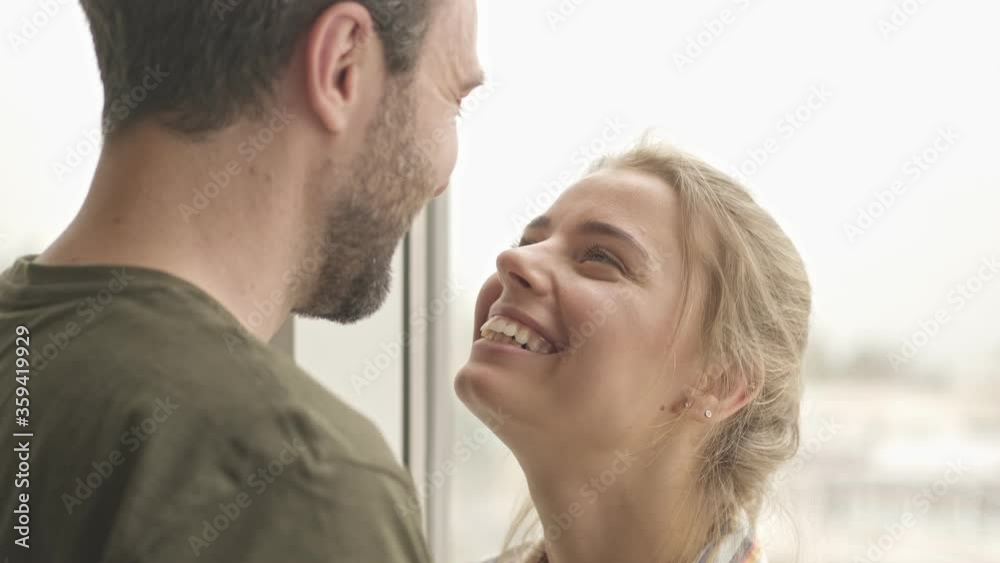 A happy young couple in love are hugging and kissing while standing near window at home A happy young couple in love are hugging and kissing while standing near window at home