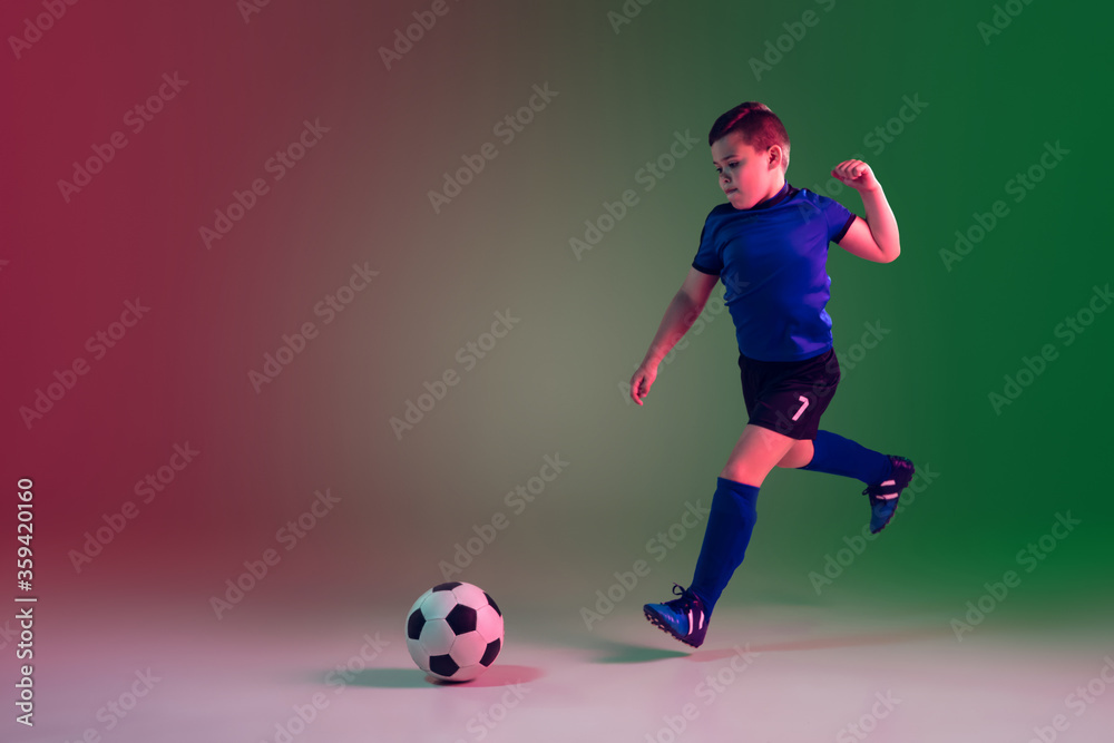 Childhood. Teen male football or soccer player on gradient background in neon light. Caucasian boy training, practicing on the run, in jump. Concept of sport, competition, winning, motion, action.