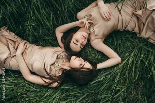 Beautiful young women twin sisters with long hair in a beige dress with chamomile flowers lie in the grass. Feelings, hugs.