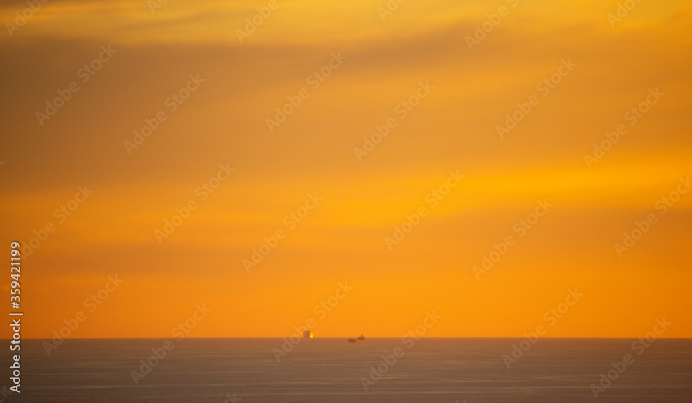 Dramatic golden sunset over ocean with cargo ships in the distance.