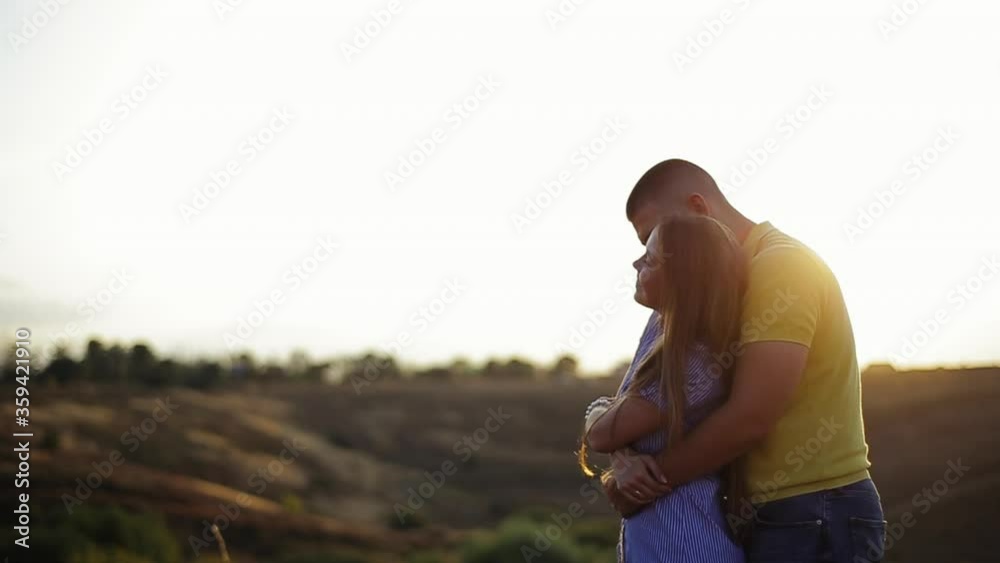 Tall young bearded guy in polo shirt hugs behind pretty girl in striped dress and hold hands in field against defocused background of sunset in evening. Romantic date in nature outside couple in love Tall young bearded guy in polo shirt hugs behind pretty girl in striped dress and hold hands in field against defocused background of sunset in evening. Romantic date in nature outside couple in love