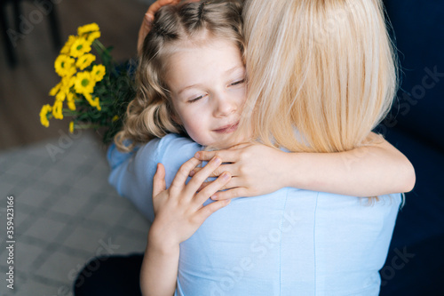 Cute mother and her adorable little daughter are hugging at the home in cozy living room. Loving daughter gives bouquet flowers to her mother.Concept of Happy Mothers Day.