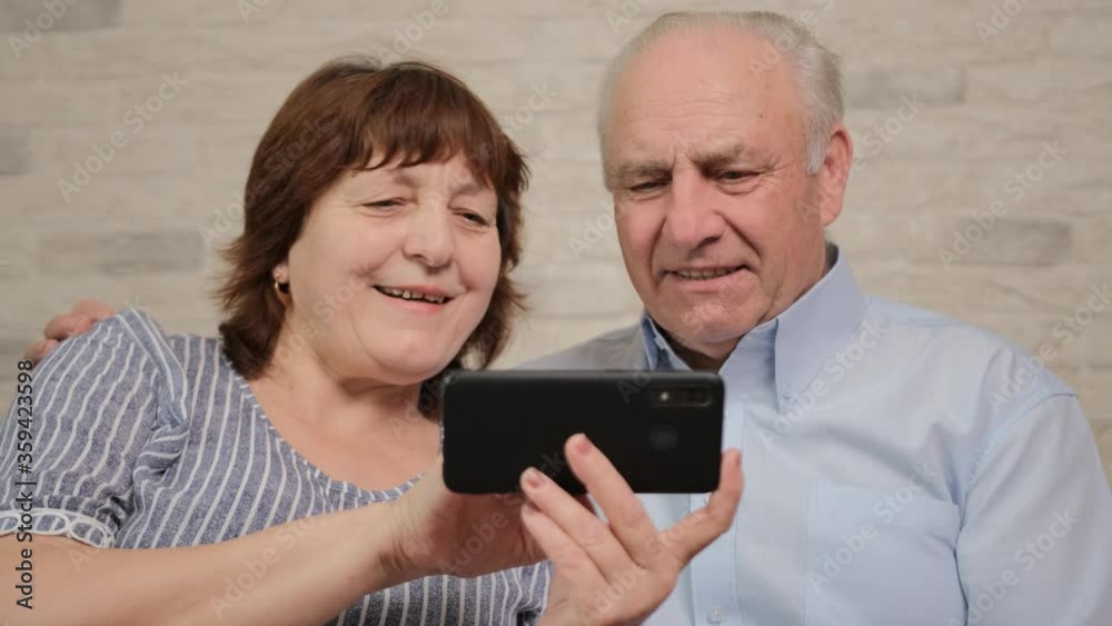 Grey-haired pensioner is helping by his aged wife with new smartphone app, hugging. Happy mature family spouse using cell phone together at home, surfing internet, sitting on couch. Grey-haired pensioner is helping by his aged wife with new smartphone app, hugging. Happy mature family spouse using cell phone together at home, surfing internet, sitting on couch.