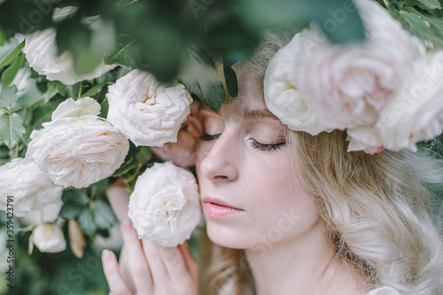  Beautiful blonde young woman enjoying smell in a rose garden. Beautiful big roses flowers. Dreaming young model between flowers