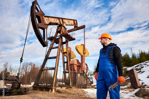 Horizontal snapshot of oil worker in orange helmet during his working day in an oilfield, oil engineer holding a pipe wrench looking at a pump jack behind him. Concept of oil and gas industry