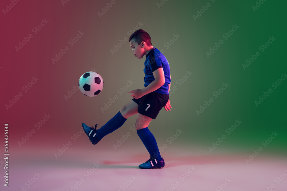 Unstoppable. Teen male football or soccer player on gradient background in neon light. Caucasian boy training, practicing on the run, in jump. Concept of sport, competition, winning, motion, action.