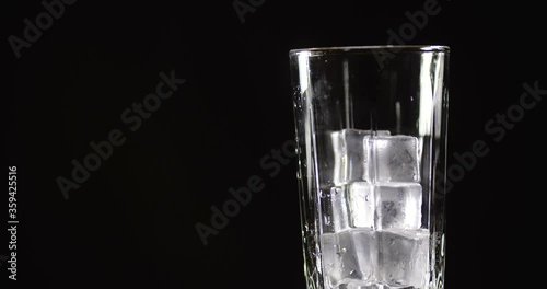 Cola with ice cubes. Side view of rotating glass in which cold carbonated brown sweet drink is poured on black background isolated with free copy space. Slow Motion 50 FPS 4k