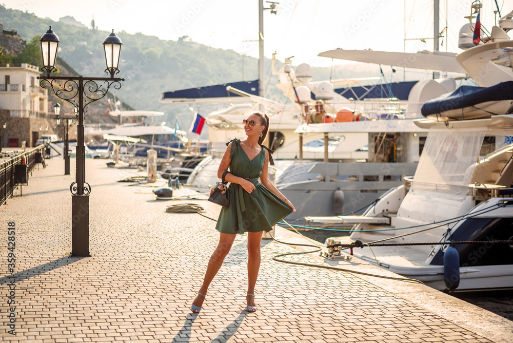 A beautiful woman in a fashionable summer dress and stylish accessories walks in the summer along the promenade next to expensive yachts