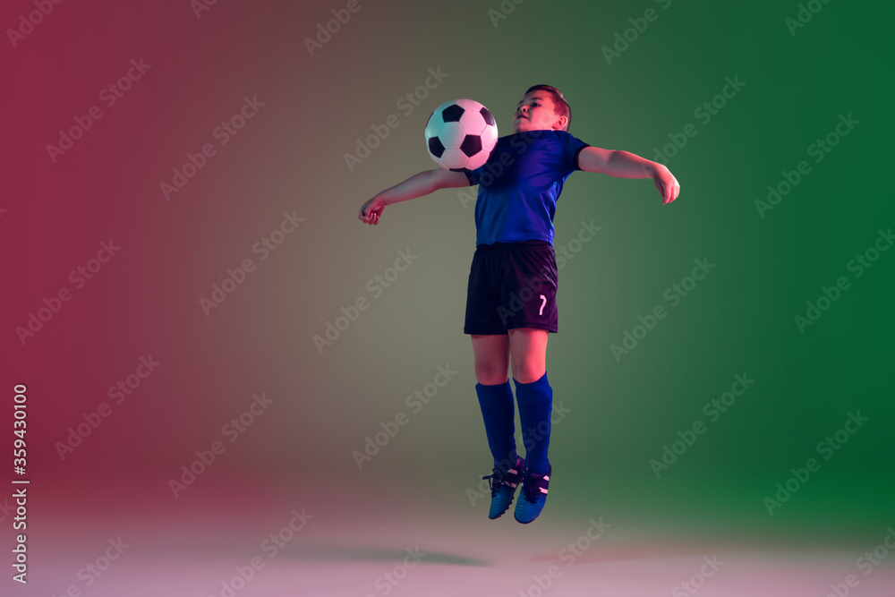 Emotions. Teen male football or soccer player on gradient background in neon light. Caucasian boy training, practicing on the run, in jump. Concept of sport, competition, winning, motion, action.