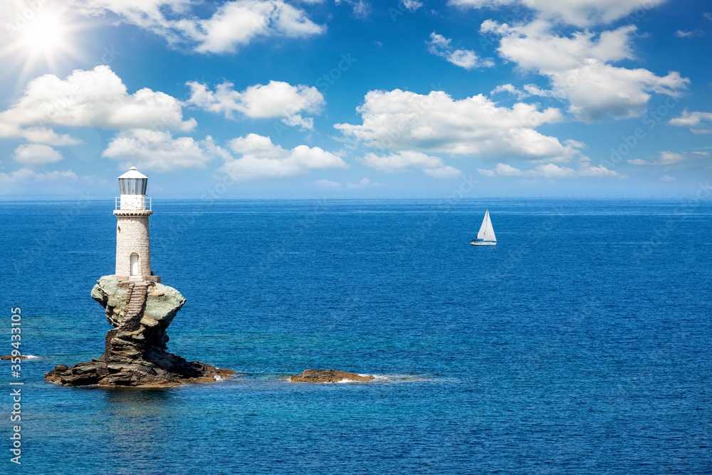 A lighthouse on a steep rock in calm, blue sea with a passing by sailing boat at the horizon, Andros, Cyclades, Greece