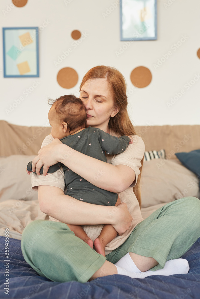 Warm-toned full length portrait of happy adult mother embracing mixed-race baby while sitting on bed at home, copy space