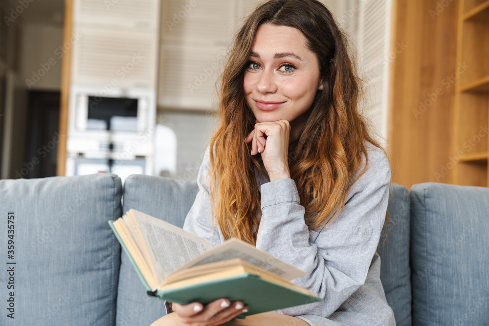 Photo of cheerful woman smiling and reading book while sitting on couch Photo of cheerful woman smiling and reading book while sitting on couch