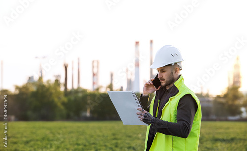 Young engineer with smartphone standing outdoors by oil refinery. Copy space.