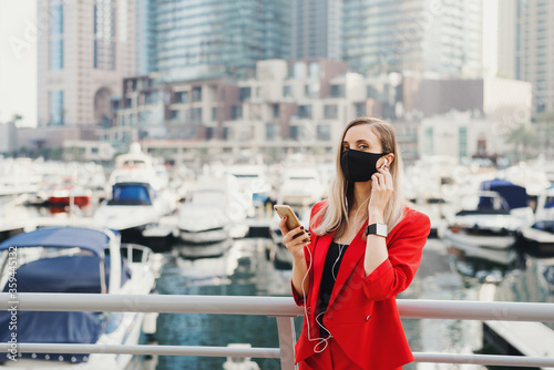 Young european businesswoman with blond hair wearing red suit and reusable protective face mask standing near yachts and skyscrapers and making a voice call via earphones. New normal  © VeronikaSmirnaya