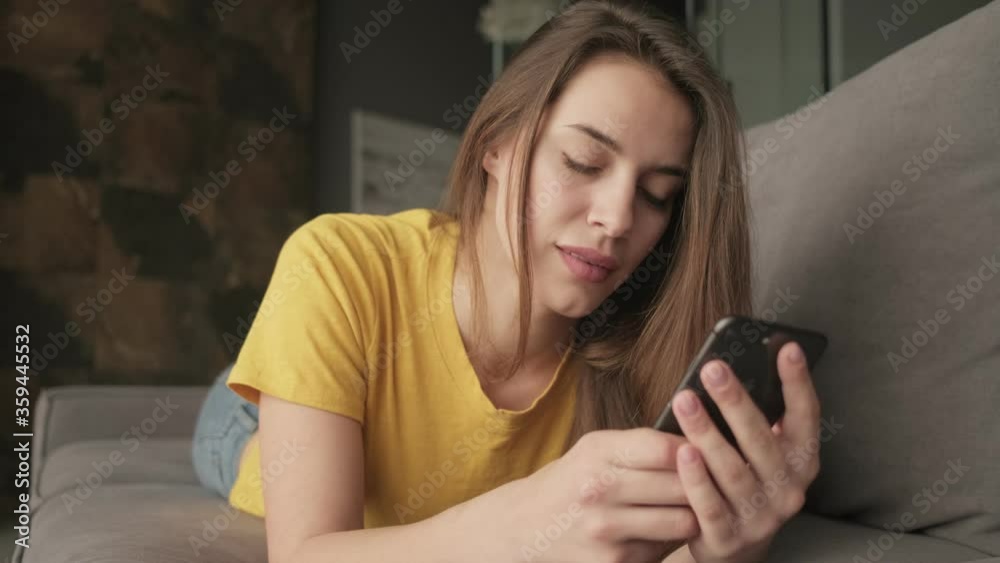A beautiful relaxed young woman is typing on her smartphone while lying on the sofa at home A beautiful relaxed young woman is typing on her smartphone while lying on the sofa at home