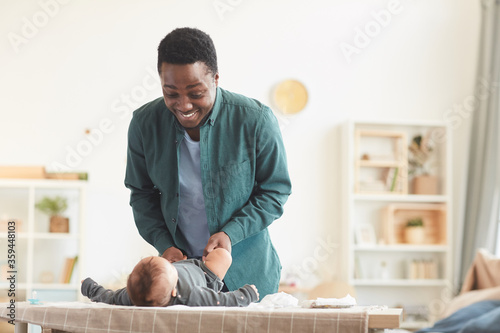 Warm-toned portrait of loving African-American father playing with cute baby boy while dressing him on changing table at home, copy space