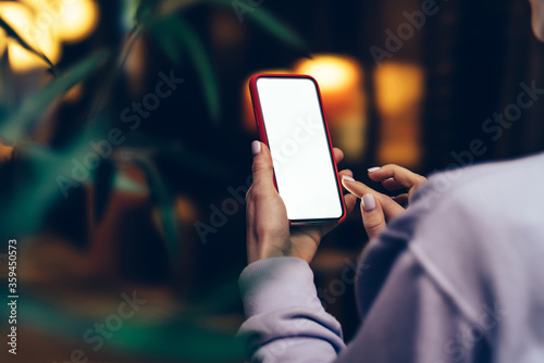 Rear view from shoulder of female person touching with finger copy space screen of modern smartphone standing outdoors near bokeh lights background. Woman using mobile phone applications with internet