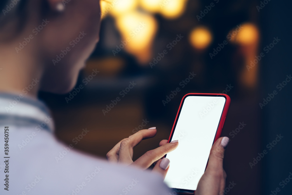 Back view from woman's shoulder of touching with finger lighted screen of smartphone device near copy space area with bokeh night lights. Texting message on mobile phone gadget with blank display