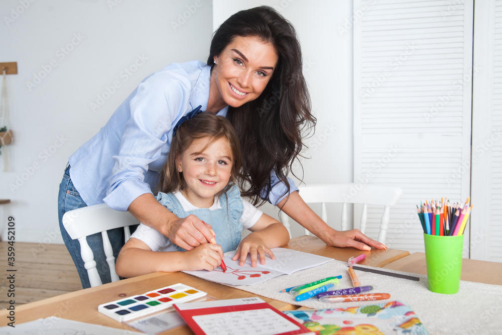 Smiling mother and daughter preparing for lessons and draws at the table with pencils and paints. Parent and pupil of preschool. First day of fall autumn. Girl from elementary class, back to school Smiling mother and daughter preparing for lessons and draws at the table with pencils and paints. Parent and pupil of preschool. First day of fall autumn. Girl from elementary class, back to school